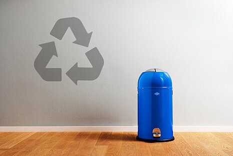 Zu sehen ist ein blauer Mülleimer rechts im Bild und ein Recycling-Symbol links im Bild