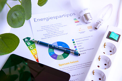 Zu sehen ist ein Blatt Papier mit der Aufschrift "Energiespartipps", eine Steckerleiste und Glühbirnen